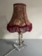 La Lampe pied en verre vintage 1940 présente un abat-jour à motifs floraux avec des franges rouges et un socle en verre clair à facettes, idéal pour ajouter un charme intemporel à n'importe quelle pièce. Brocante 93
