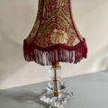 La Lampe pied en verre vintage 1940, dotée d'un abat-jour floral rouge et vert frangé et d'une base en verre transparent, repose sur une surface grise et apporte un charme vintage à tout espace. Brocante 93