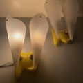 Paire d'appliques vintage 1960 en forme d'oiseaux, dotées d'abat-jours blancs et de bases jaunes, diffusant une lumière douce sur une surface grise. Brocante 93