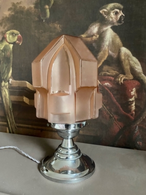 La Lampe Art Déco 1930, une lampe géométrique rose avec une base chromée, est exposée devant un tableau représentant un singe et un perroquet. Brocante 93