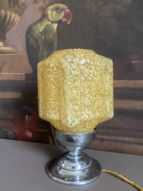 Lampe en verre 1950 avec un abat-jour en verre jaune texturé et une base chromée, placée devant un tableau de perroquets. Brocante 93