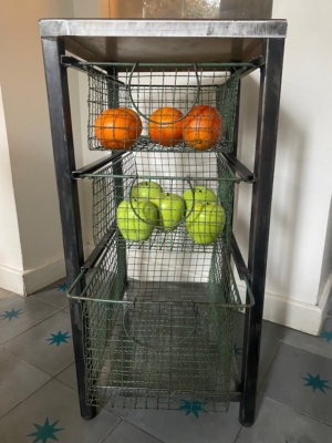 Une étagère métallique avec des paniers en fil de fer contient des oranges sur l'étagère du haut et des pommes vertes sur l'étagère du milieu. Brocante 93