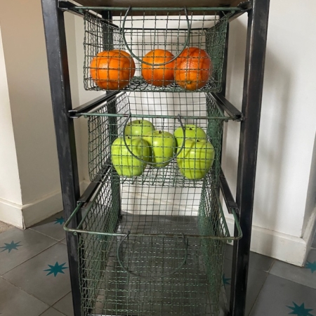 Une étagère métallique avec des paniers en fil de fer contient des oranges sur l'étagère du haut et des pommes vertes sur l'étagère du milieu. Brocante 93