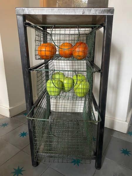 Une étagère métallique avec des paniers en fil de fer contient des oranges sur l'étagère du haut et des pommes vertes sur l'étagère du milieu. Brocante 93