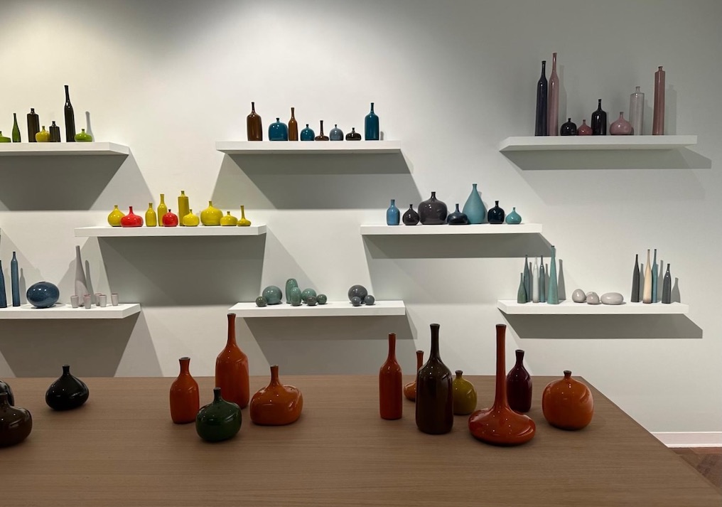 Des vases colorés en verre Carouche de différentes formes sont présentés sur des étagères murales blanches et une table en bois. Brocante 93