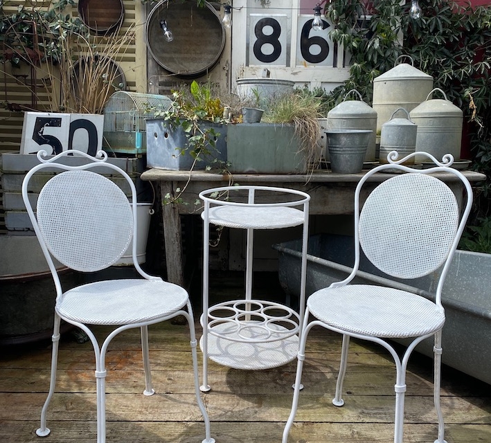 Deux chaises blanches en métal et une petite table sur un patio en bois, entouré de plantes et de décorations de jardin vintage. Brocante 93