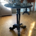 La Guéridon Napoléon III, une petite table ronde noire, est surmontée d'objets décoratifs en verre et posée sur un plancher en bois dans une pièce ensoleillée. Brocante 93