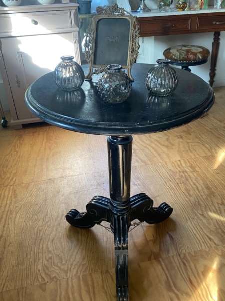 Guéridon Napoléon III : Petite table ronde noire antique avec trois vases en verre et un cadre orné, posée sur un plancher en bois. Brocante 93
