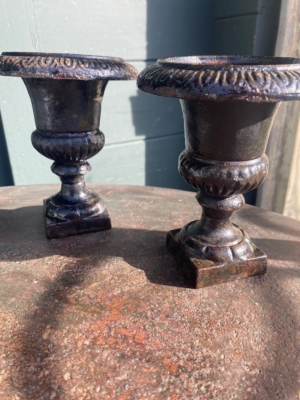 Une paire de vases Médicis 1940, petits et ornés en métal noir, repose sur une surface métallique rouillée sous la lumière du soleil. Brocante 93