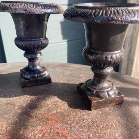 Une paire de vases Médicis 1940, petits et ornés en métal noir, repose sur une surface métallique rouillée sous la lumière du soleil. Brocante 93
