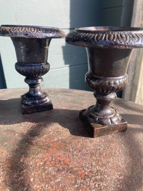 Une paire de vases Médicis 1940, petits et ornés en métal noir, repose sur une surface métallique rouillée sous la lumière du soleil. Brocante 93