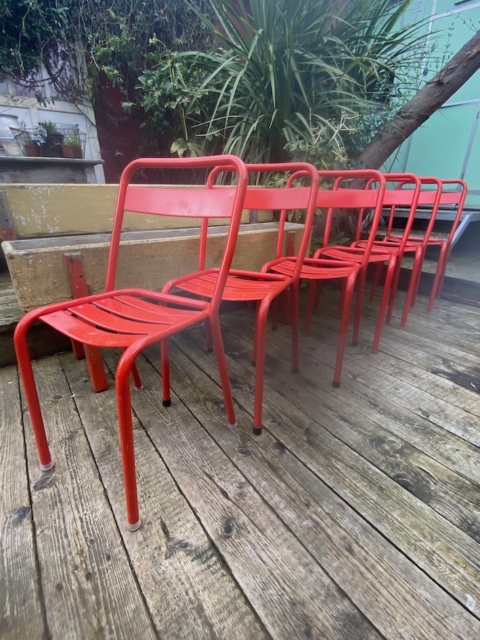 Six chaises du lot de chaises métal 1960 sont alignées sur un deck en bois, avec des plantes et un banc à l'arrière-plan. Brocante 93