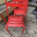 Quatre chaises métalliques rouges sont alignées sur une terrasse en bois, à côté d'un banc et de quelques plantes en pot. Brocante 93