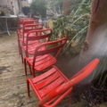 Cinq chaises métalliques rouges sont alignées sur une terrasse en bois, entourée de plantes vertes et de mobilier d'extérieur. Brocante 93