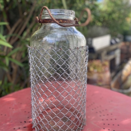 Une Bombonne grillagée 1950 est posée à l'extérieur sur une table rouge perforée, son pot en verre étant enfermé dans un solide couvercle en métal. Brocante 93