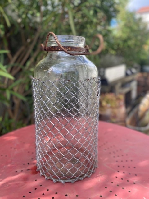 Une Bombonne grillagée 1950 est posée à l'extérieur sur une table rouge perforée, son pot en verre étant enfermé dans un solide couvercle en métal. Brocante 93