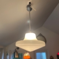La lampe suspendue Suspension verre & opaline 1940 est suspendue au plafond dans une pièce dont les fenêtres et les autres lumières sont visibles en arrière-plan. Brocante 93