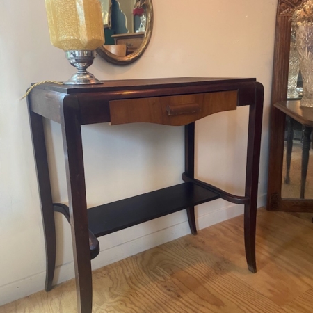 La Console Art Déco, une table console en bois foncé avec un tiroir, est stylisée avec une lampe et un miroir rond au-dessus, placés contre un mur clair et un sol en bois. Brocante 93
