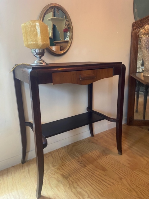 La Console Art Déco, une table console en bois foncé avec un tiroir, est stylisée avec une lampe et un miroir rond au-dessus, placés contre un mur clair et un sol en bois. Brocante 93