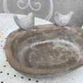 Le Bain oiseaux pierre 1960 est un bain d'oiseaux en pierre vieillie datant de 1960, avec deux petites sculptures d'oiseaux sur son rebord, présentées sur une table blanche perforée. Brocante 93