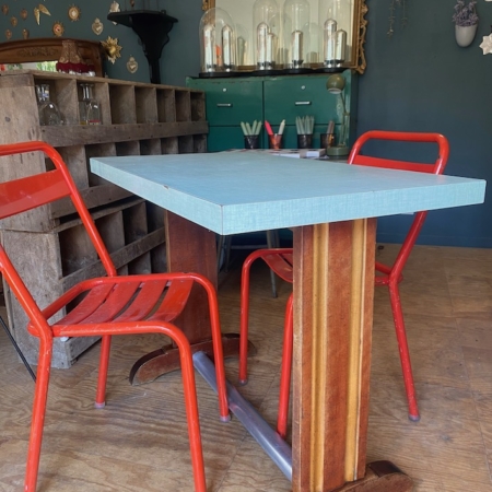 Une table bistrot Formica 1950, bleue et rectangulaire, associée à deux chaises en métal rouge, se trouve dans une pièce vivante décorée de manière éclectique dans les années 1950. Brocante 93