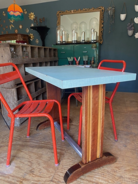 Une table bistrot Formica 1950, bleue et rectangulaire, associée à deux chaises en métal rouge, se trouve dans une pièce vivante décorée de manière éclectique dans les années 1950. Brocante 93