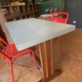 La table bistrot Formica 1950 présente un plateau rectangulaire aux tons clairs et une base en bois, associée à des chaises en métal rouge dans une pièce de style rétro des années 1950. Brocante 93