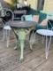 Guéridon de jardin en fonte 1940, table ronde en métal ornée, accompagnée de deux chaises métalliques blanches sur terrasse en bois. Brocante 93