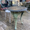 Le Guéridon de jardin en fonte 1940 présente un motif en fonte orné d'une patine verte, présenté sur une terrasse en bois à côté d'une chaise de bistrot en métal blanc. Brocante 93