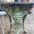 Pied de table en fonte verte provenant d'un Guéridon de jardin en fonte de 1940, présentant des figures sculptées, exposées sur une terrasse en bois. Brocante 93