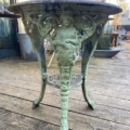 Guéridon de jardin en fonte 1940, vert patiné, avec pieds ornés de figures humaines sculptées, exposé sur terrasse en bois. Brocante 93