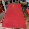 Une table de bistrot Formica rouge 1950 avec un plateau rectangulaire rouge et deux chaises assorties est posée sur un sol en bois, évoquant une ambiance rustique des années 1950. Brocante 93