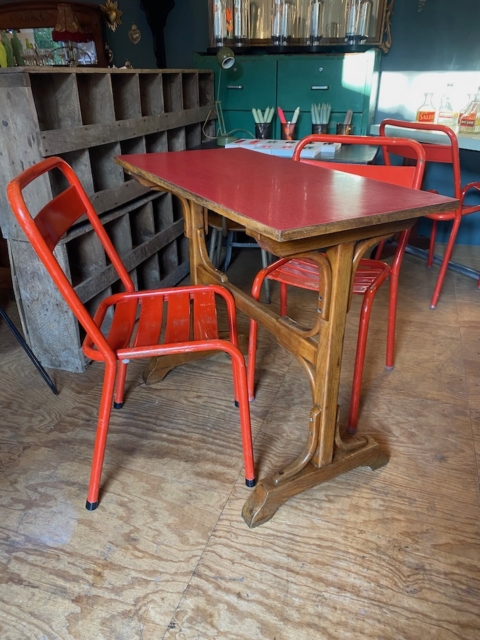 Un café confortable présente la Table de bistrot Formica rouge 1950 associée à des chaises en métal rouge, entourée d'un décor en bois chaleureux et en métal industriel. Brocante 93