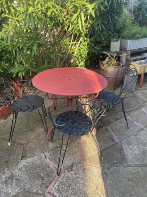 Le Salon de jardin fer forgé 1960 présente une table ronde rouge en métal et trois chaises noires, créant ainsi un ensemble chic et vintage sur un patio en pierre au milieu d'une végétation luxuriante. Brocante 93