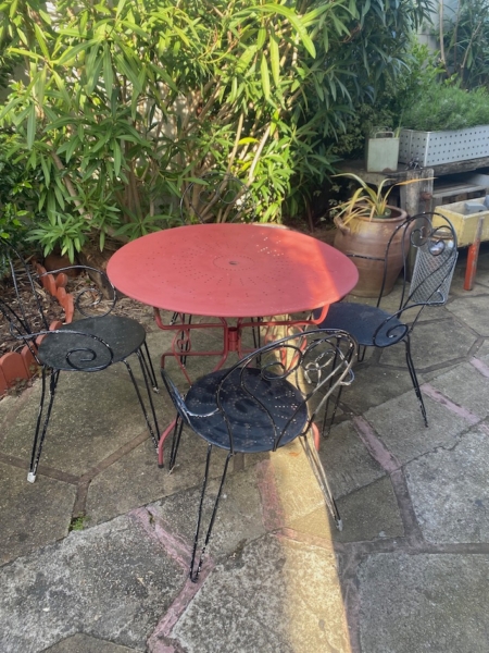Le Salon de jardin fer forgé 1960 présente une table ronde rouge en métal et trois chaises noires, créant ainsi un ensemble chic et vintage sur un patio en pierre au milieu d'une végétation luxuriante. Brocante 93
