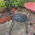 Le Salon de jardin fer forgé 1960, une chaise en métal noir avec un motif en forme de tourbillon, est associé à une table en métal rouge dans un jardin. Brocante 93