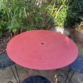 Le Salon de jardin fer forgé 1960 comprend une table ronde en métal rouge et trois chaises en fer forgé noir, parfaites pour une installation en extérieur parmi des plantes vertes et des arbustes en pot, créant un charme nostalgique des années 1960. Brocante 93