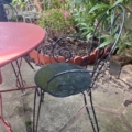 Le Salon de jardin fer forgé 1960, avec une chaise en métal noir et une table rouge, est posé sur un patio en pierre avec des plantes et un parterre de fleurs en arrière-plan. Brocante 93