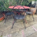 Le Salon de jardin fer forgé 1960 - un ensemble de trois chaises en métal noir et une table ronde rouge - est posé sur un patio en pierre parmi des plantes vertes et des pots, créant ainsi l'espace extérieur vintage parfait. Brocante 93