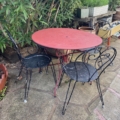 Le Salon de jardin fer forgé 1960 présente une table ronde en métal rouge et deux chaises en fer forgé noir sur un patio en pierre parmi des plantes luxuriantes, évoquant une charmante atmosphère de salon de jardin des années 1960. Brocante 93
