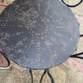 Le Salon de jardin fer forgé 1960 est une table ronde en métal noir usé par les intempéries, dont la surface présente des taches de rouille et de petits trous, posée sur un dallage en pierre. Brocante 93