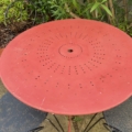 Le Salon de jardin fer forgé 1960 présente une table ronde en métal rouge et deux chaises noires, créant un charmant look vintage dans un jardin luxuriant entouré de verdure. Brocante 93