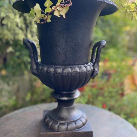 Le Vase Médicis XXème siècle, une urne noire ornementale avec des vignes vertes, se pose sur une table de jardin ronde à l'extérieur, apportant une élégance XXème siècle à votre espace. Brocante 93