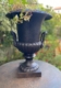 Le Vase Médicis XXème siècle, une urne noire ornementale avec des vignes vertes, se pose sur une table de jardin ronde à l'extérieur, apportant une élégance XXème siècle à votre espace. Brocante 93