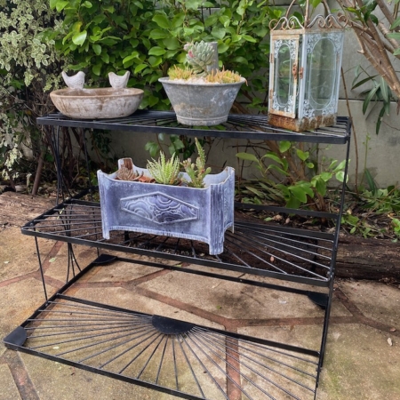 Le Présentoir à plantes 1970 contient des plantes grasses en pot et une lanterne en verre à l'extérieur, au milieu d'un feuillage vert luxuriant. Brocante 93