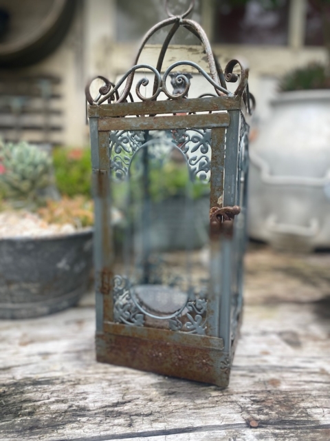 La Lanterne de jardin 1940, ornée de détails et de panneaux de verre, repose sur une surface en bois, évoquant une ambiance de jardin classique. Brocante 93