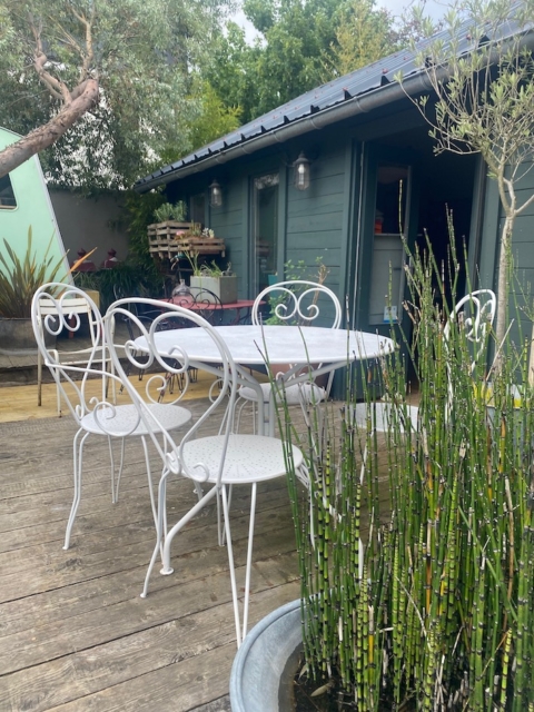 Le Salon de jardin en fer forgé 1950 ajoute un charme vintage à une terrasse en bois près d'un abri de verdure, entouré de plantes luxuriantes. Brocante 93