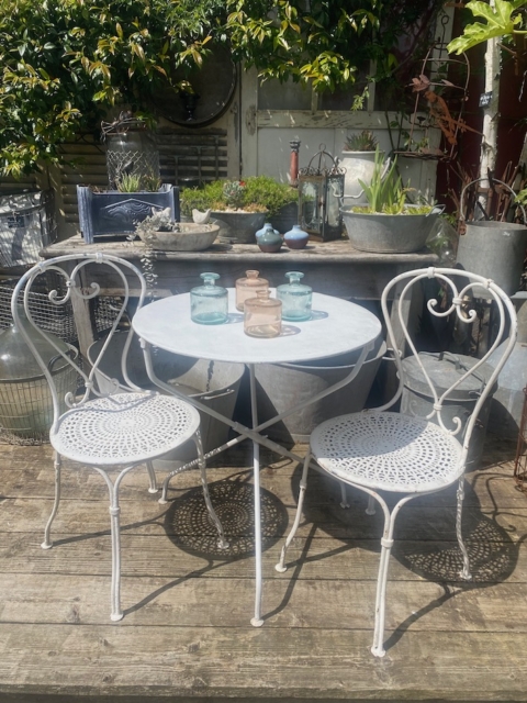 L'ensemble Guéridon & 2 chaises en fer forgé 1950, disposé sur une terrasse en bois dans un jardin ensoleillé avec des jarres, évoque le charme intemporel du mobilier vintage en fer forgé. Brocante 93