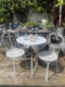 L'ensemble Guéridon & 2 chaises en fer forgé 1950, disposé sur une terrasse en bois dans un jardin ensoleillé avec des jarres, évoque le charme intemporel du mobilier vintage en fer forgé. Brocante 93
