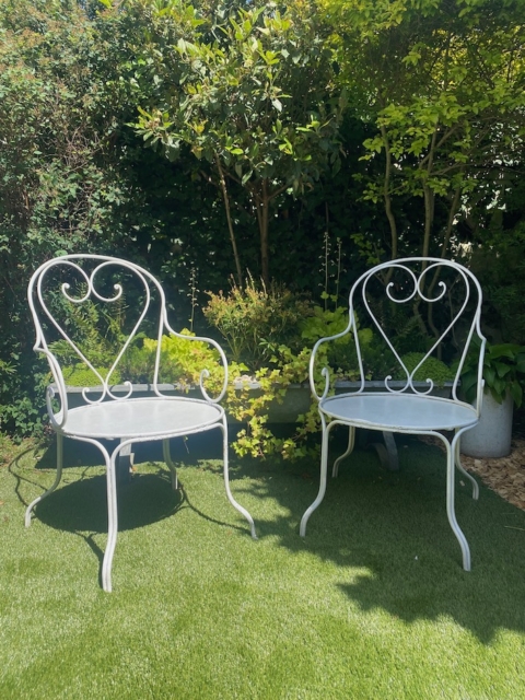 Une paire de fauteuils fer forgé 1950 est placée sur l'herbe verte d'un jardin ensoleillé, entourée de plantes et de buissons luxuriants. Brocante 93